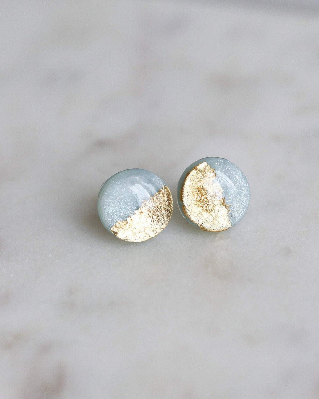 Round minimalist stud earrings with gold and light blue accents on a marble surface