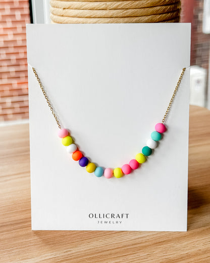 Colorful beaded necklace on a white card with 'Ollicraft Jewelry' branding, placed on a wooden surface.