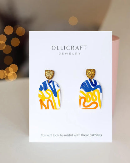 Colorful abstract-patterned earrings on Ollicraft branded card with blurred lights in the background