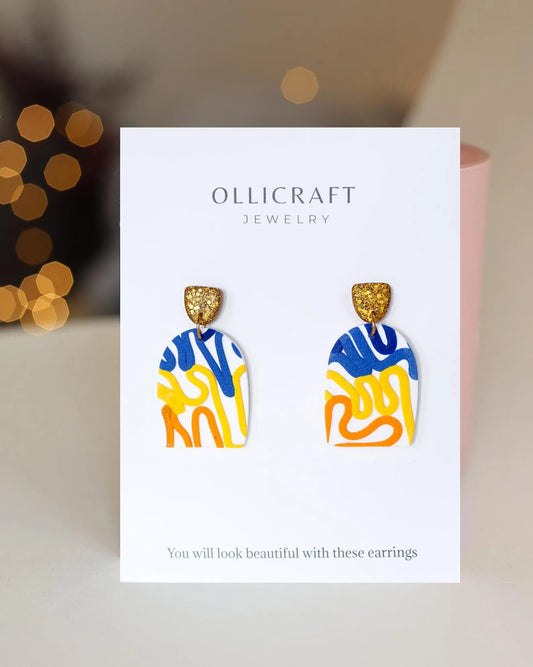 Colorful abstract-patterned earrings on Ollicraft branded card with blurred lights in the background