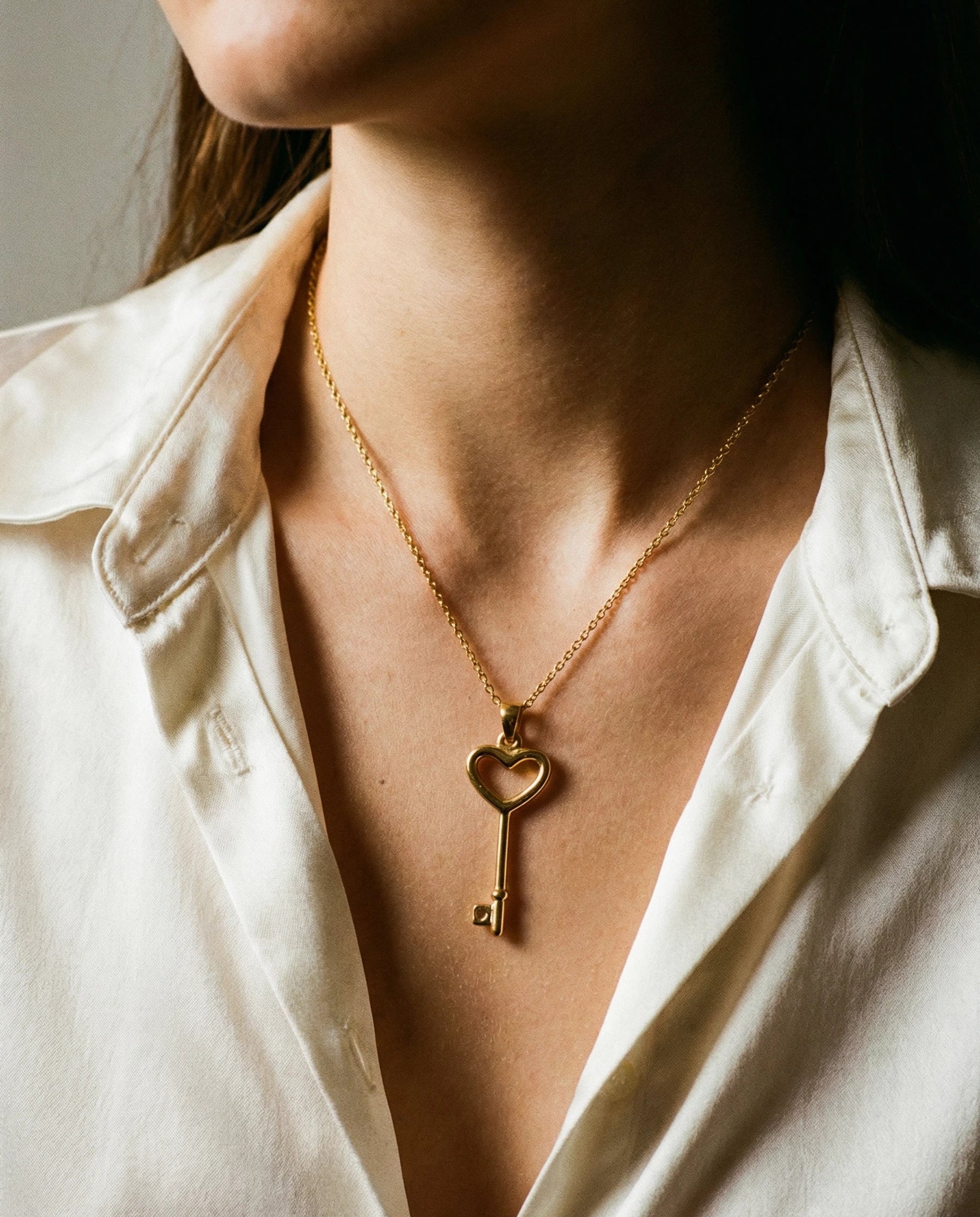 Gold necklace with a heart-shaped key pendant worn by a person in a white shirt.